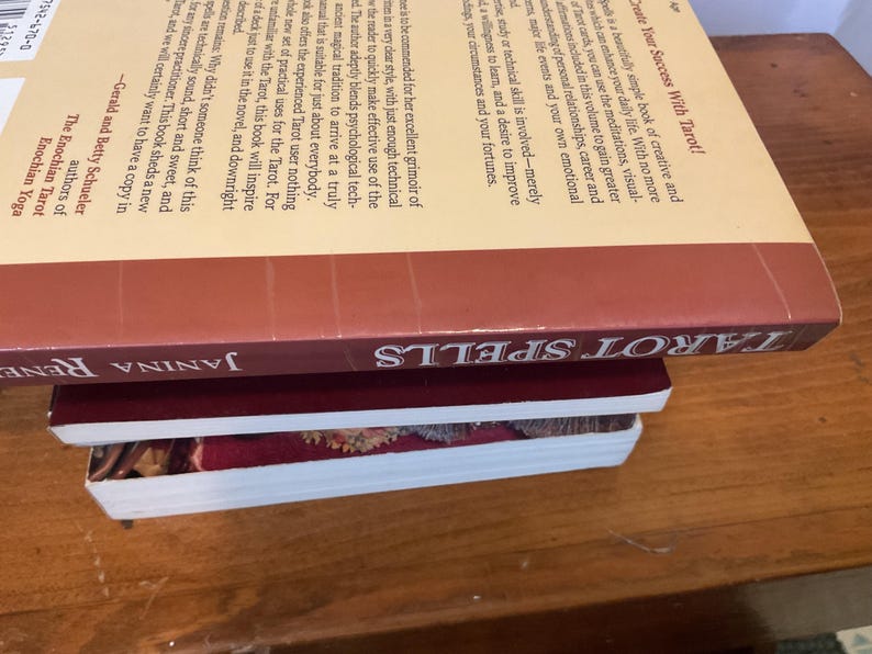 May include: A stack of three books. The top book is titled Tarot Spells by Janina Renee, featuring a cream-colored back cover with black text and a maroon spine. Below it is a thin maroon book, and the bottom book has a white spine and cover.