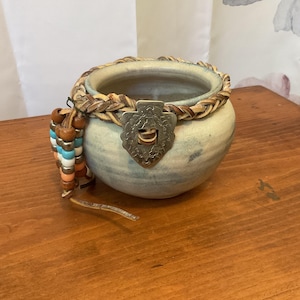 May include: A small, round, ceramic pot with a braided leather trim and a decorative metal clasp. A beaded strand with wooden and colorful beads hangs from the side. The pot has a blue and white mottled glaze.
