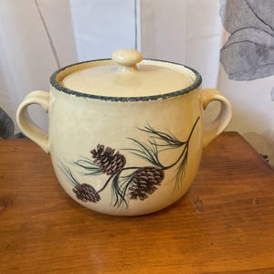 May include: Cream-colored ceramic pot with lid, featuring a pine cone and needle design. The pot has two handles and a green trim around the rim. The lid has a small knob. The pot is sitting on a wooden surface.