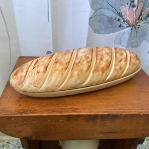 May include: A ceramic bread-shaped serving dish with a light brown and cream color scheme. The dish is placed on a wooden table. The bread-shaped dish has vertical grooves and is designed to resemble a loaf of bread.