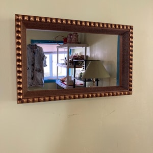 May include: A gold-toned framed mirror with a beveled edge hangs on a light yellow wall. The mirror reflects a room with a shelf unit, a lamp, and a window with a blue trim.