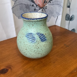 May include: A speckled, light green ceramic vase with a rounded body and a blue rim. Two blue, stylized wing-like designs are painted on the front. The vase is displayed on a wooden surface.
