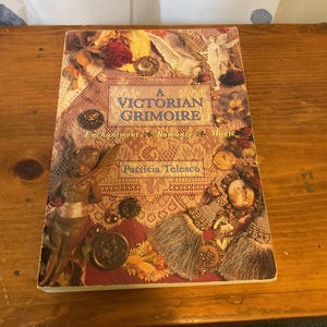 May include: A paperback book titled A Victorian Grimoire by Patricia Telesco. The cover features a collage of lace, a winged cherub, dried roses, tassels, and vintage-style brooches in warm tones of gold, cream, and deep red.