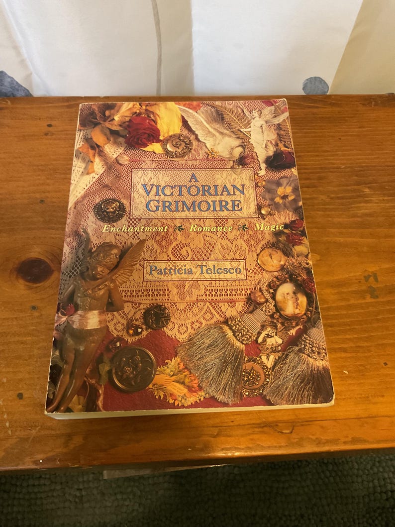 May include: A paperback book titled A Victorian Grimoire by Patricia Telesco. The cover features a collage of lace, a winged cherub, dried roses, tassels, and vintage-style brooches in warm tones of gold, cream, and deep red.