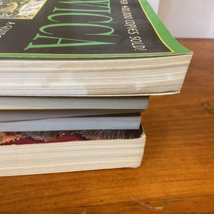 May include: A stack of three books with visible page edges. The top book has a black cover with green text reading WICCA and white text stating A GUIDE FOR THE SOLITARY PRACTITIONER and OVER 400,000 COPIES SOLD. The bottom book has a dark red cover.