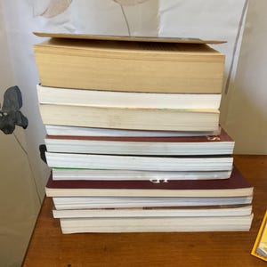 May include: A vertical stack of eleven assorted books with varying spine colors including tan, white, and dark maroon. Beside the stack lies a rectangular yellow bookmark featuring the text "BOOKS FALL OPEN YOU FALL IN" in black lettering.
