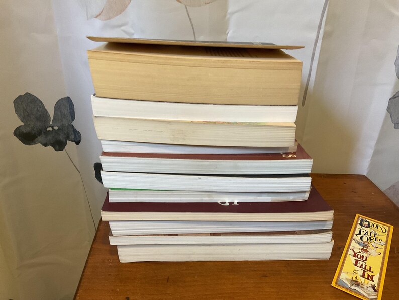 May include: A vertical stack of eleven assorted books with varying spine colors including tan, white, and dark maroon. Beside the stack lies a rectangular yellow bookmark featuring the text "BOOKS FALL OPEN YOU FALL IN" in black lettering.