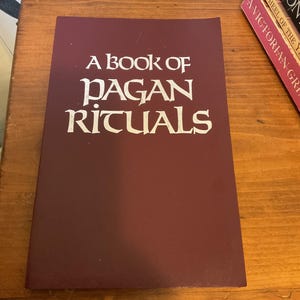 May include: A rectangular book with a dark maroon cover featuring the white text A BOOK OF PAGAN RITUALS centered on the front. The book is positioned flat, with the title clearly printed in a stylized, serif font against the solid background.