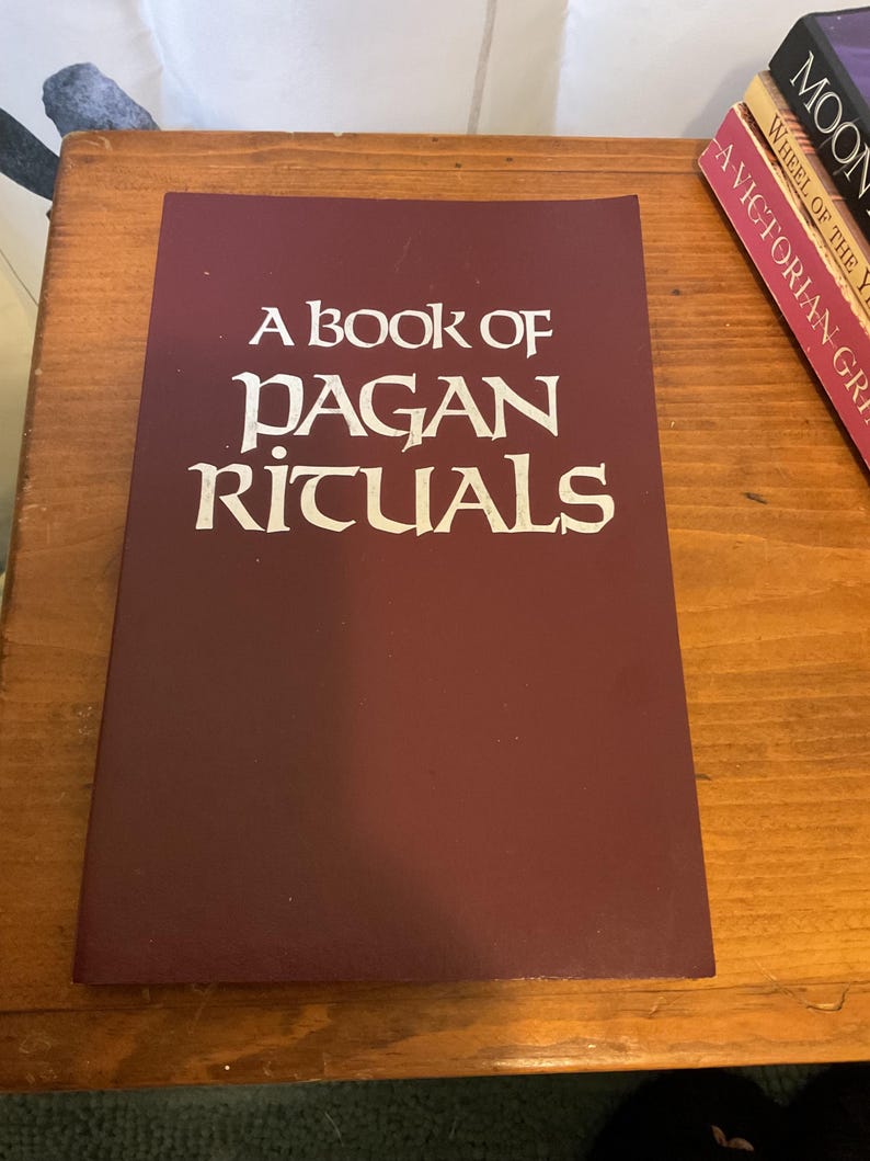 May include: A rectangular book with a dark maroon cover featuring the white text A BOOK OF PAGAN RITUALS centered on the front. The book is positioned flat, with the title clearly printed in a stylized, serif font against the solid background.
