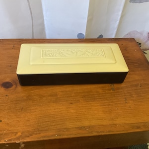 May include: A rectangular, two-tone box with a cream-colored lid and a dark brown base. The lid features a raised, decorative design. The box is resting on a wooden surface, suggesting a vintage or antique aesthetic.