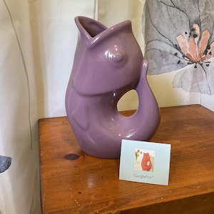 May include: A glossy, light purple ceramic pitcher shaped like a fish with an open mouth and a curved handle. Next to the pitcher is a small, light blue rectangular card featuring an illustration of a red fish pitcher and the text GurglePot.