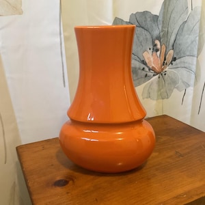 May include: A vibrant orange vase with a bulbous base and a flared top. The glossy ceramic vase sits on a wooden surface, with a floral patterned curtain in the background. The vase has a smooth, rounded shape.