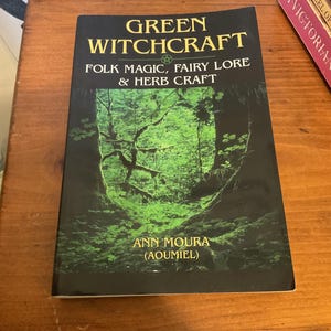 May include: A paperback book titled Green Witchcraft: Folk Magic, Fairy Lore & Herb Craft by Ann Moura (Aoumiel). The cover features a dark forest scene with vibrant green foliage and trees, with yellow text against a black background.