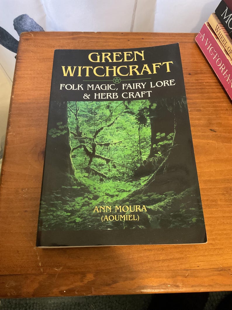 May include: A paperback book titled Green Witchcraft: Folk Magic, Fairy Lore & Herb Craft by Ann Moura (Aoumiel). The cover features a dark forest scene with vibrant green foliage and trees, with yellow text against a black background.