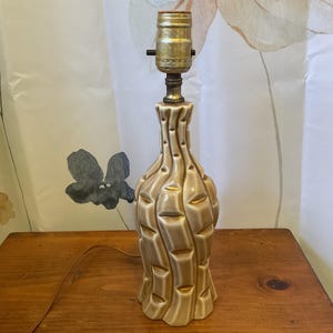 May include: A vintage beige ceramic table lamp with a textured, geometric design. The lamp has a brass-colored socket and a brown electrical cord. The lamp is sitting on a wooden surface.