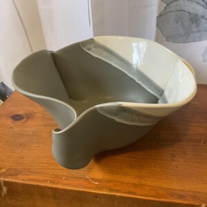 May include: A ceramic bowl with a unique, asymmetrical shape. The bowl is primarily gray, with a section of white and gray accents. The rim has a wavy design. The bowl is sitting on a wooden surface.