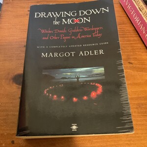 May include: A paperback book titled Drawing Down the Moon by Margot Adler with a black cover. The cover features white and red text, a subtitle about Witches and Druids, and a central image of a person standing in a circle of glowing red candles.