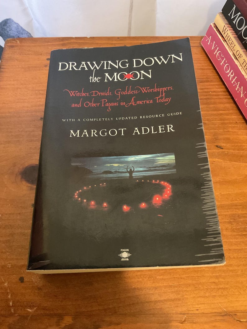 May include: A paperback book titled Drawing Down the Moon by Margot Adler with a black cover. The cover features white and red text, a subtitle about Witches and Druids, and a central image of a person standing in a circle of glowing red candles.