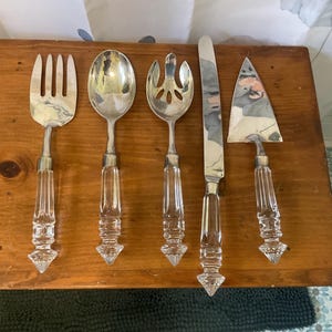 May include: A set of five serving utensils, including a fork, spoon, slotted spoon, knife, and cake server, arranged on a wooden surface. The utensils have silver-colored heads and clear, faceted handles. The cake server has a triangular blade.