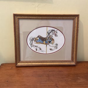 May include: Framed artwork featuring a carousel horse illustration. The horse is white with a blue and brown saddle, set against a neutral background within an oval frame. The frame is gold-toned, adding a touch of elegance.