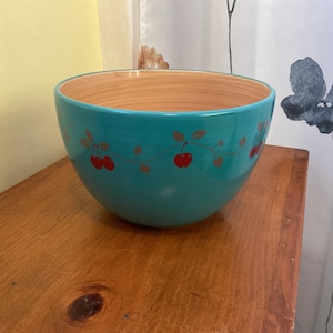 May include: A teal ceramic bowl with a light brown interior. The exterior is decorated with a cherry vine design in red and gold. The bowl sits on a wooden surface, suggesting a kitchen or dining setting.