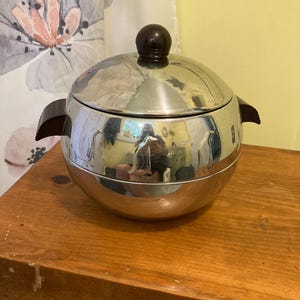 May include: A spherical silver-toned metal ice bucket featuring a polished finish and embossed penguin motifs around the center. The container includes a matching lid with a dark brown spherical knob and two dark brown side handles for lifting.