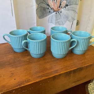 May include: Six turquoise ceramic mugs with vertical ribbed detailing. Each mug has a rounded base and a curved handle. The mugs are arranged on a wooden surface, suggesting a kitchen or dining setting.