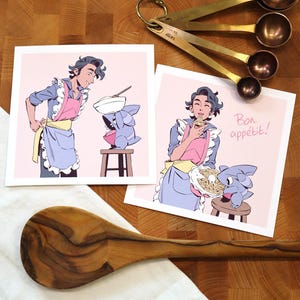 Puede incluir: Dos impresiones artísticas cuadradas que representan a un chef de dibujos animados y una criatura parecida a un tiburón cocinando. El chef lleva un delantal rosa y un delantal azul. Una impresión dice "Bon appétit!" en cursiva. También se ven cucharas medidoras y una cuchara de madera.