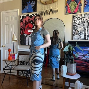 May include: A woman wearing a denim overall dress with a large eye painted on the front. The dress is a dark blue denim with a light blue wash. The eye is painted in black and white with a white outline. The woman is wearing white boots and has long brown hair. She is standing in a room with a mirror behind her. The room is decorated with colorful paintings on the walls.