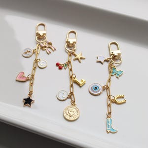 May include: Three gold keychains with charms. The first keychain has a pink heart, a black star, a white cross, a white letter C, a pink bow, and a gold coin with an eagle design. The second keychain has a red cherry, a white smiley face, a gold letter L, a gold starfish, and a gold coin with an eagle design. The third keychain has a blue eye, a white star, a blue butterfly, a gold horseshoe, a blue cowboy boot, and a gold chain.