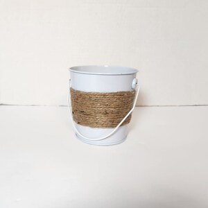 Op de afbeelding: Een kleine witte metalen emmer met een bruin touw handvat. De emmer is perfect voor het bewaren van kleine voorwerpen of planten.