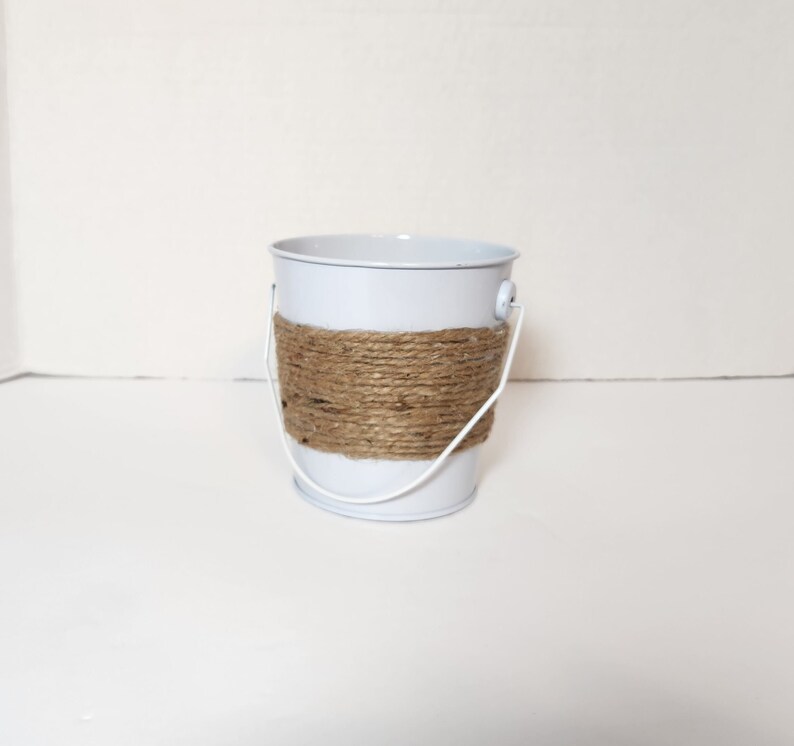 Op de afbeelding: Een kleine witte metalen emmer met een bruin touw handvat. De emmer is perfect voor het bewaren van kleine voorwerpen of planten.