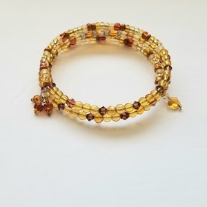 May include: A multi-strand wrap bracelet constructed from translucent amber, gold, and dark brown glass beads. The design features a spiral coil structure with small dangling bead charms attached at the ends, showcasing a mix of round and faceted bead shapes.