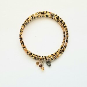 Könnte beinhalten: Ein mehrsträngiges Wickelarmband aus kleinen zylindrischen Perlen in Gold-, Bronze- und Schwarztönen. Das Armband ist mit drei hängenden Anhängern versehen, darunter zwei kleine facettierte bernsteinfarbene Kristalle und ein silberfarbenes Blatt.