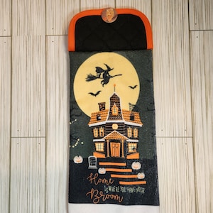 May include: Halloween-themed kitchen towel with a black and orange top and a decorative button. The main design features a haunted house, a witch flying on a broom, and the text "Home is where you hang your broom."