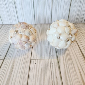 May include: Two white and beige decorative spheres made of seashells. The spheres are decorated with small white and gold beads.