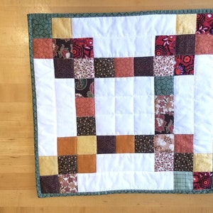 May include: A square quilted wall hanging featuring a central white block surrounded by a patchwork border of small squares in shades of brown, orange, red, and yellow, finished with a dark green patterned fabric binding along the edges.