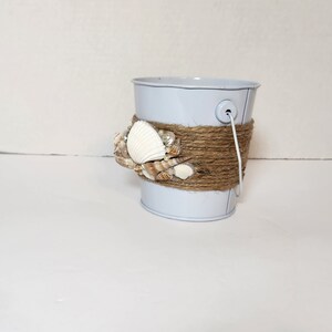Op de afbeelding: Een kleine witte metalen emmer met een bruin touw handvat en schelpen die aan de bovenkant zijn gelijmd.