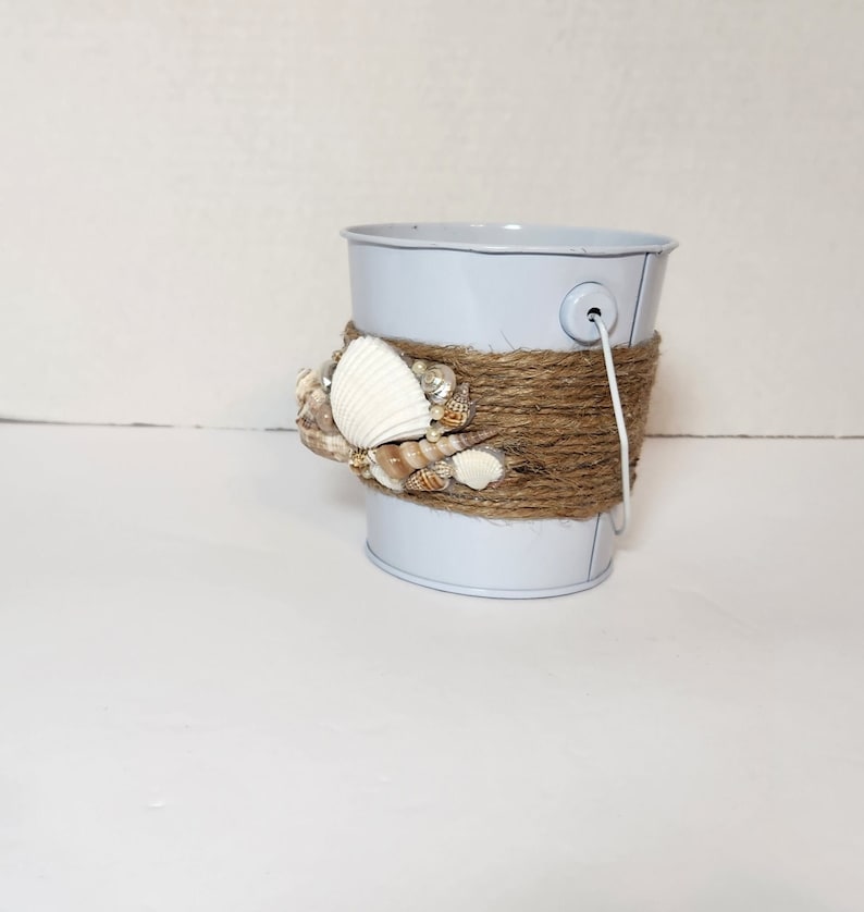 Op de afbeelding: Een kleine witte metalen emmer met een bruin touw handvat en schelpen die aan de bovenkant zijn gelijmd.