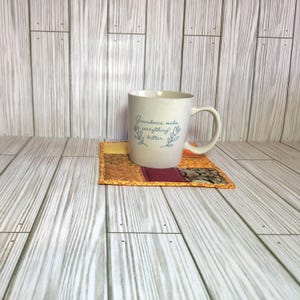 May include: A speckled off-white ceramic mug with a handle and the blue cursive text "Grandmas make everything better" flanked by leaf illustrations, resting on a square fabric coaster featuring a patchwork of orange, gold, and maroon patterns.