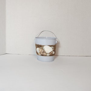 Op de afbeelding: Een kleine witte metalen emmer met een bruin touw handvat en een decoratieve band van schelpen en touw rond het midden.