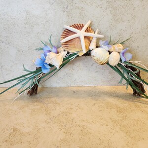 May include: A decorative arch featuring seashells, a starfish, and blue flowers. The arch is made of a brown twig base with green and white accents. Perfect for coastal or beach-themed decor.