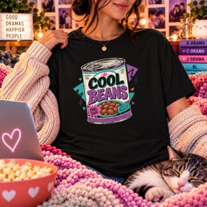 Puede incluir: Una camiseta negra con un gráfico de una lata de conserva con el texto COOL BEANS en letras moradas y negras. El diseño incluye un pequeño icono de cara sonriente rosa, frijoles marrones dentro de la lata y formas geométricas.