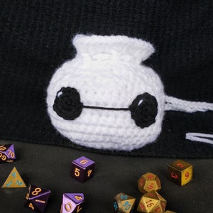 May include: A white crocheted bag with black eyes and a white drawstring. The bag is sitting on a black surface with a variety of metal dice in different colors.