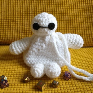 May include: White crocheted plush toy shaped like a robot with black eyes and a white drawstring bag. The toy is sitting on a yellow surface with multiple gold and purple dice.