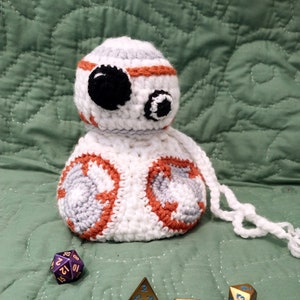May include: A white crocheted BB-8 character with orange and gray accents. The character is holding a purple 20-sided die and there are several gold dice scattered around it on a green quilted surface.