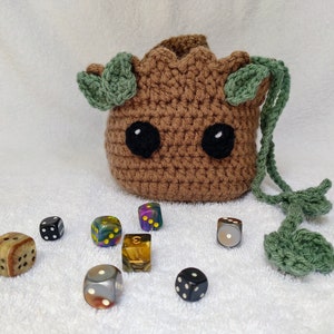May include: A brown crocheted dice bag shaped like a character with green leaves and black eyes. The bag is sitting on a white surface with eight colorful dice scattered around it.