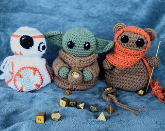 May include: Three crocheted plush toys, a white and orange BB-8, a green and brown Yoda, and a brown bear wearing an orange scarf, are arranged on a blue fuzzy surface with gold dice scattered around them.