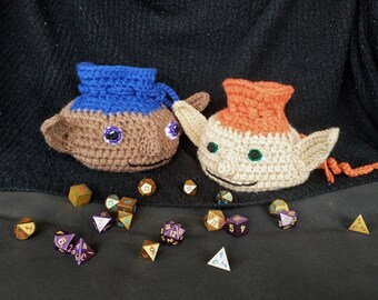 May include: Two crocheted dice bags, one blue and one orange, with brown faces and green eyes, are sitting on a black surface with a variety of dice scattered around them.