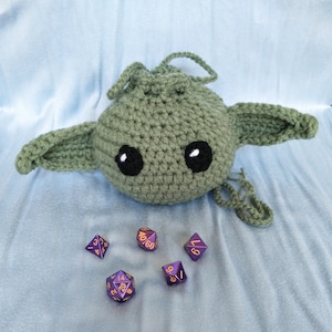Op de afbeelding: Een groene, gebreide, door Yoda geïnspireerde trekkoordtas met grote zwarte ogen. Vijf paarse en gouden dobbelstenen liggen verspreid op een lichtblauw oppervlak.